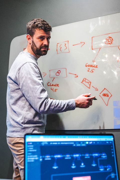 Jacob Lynov Mindak står ved et whiteboard på en mørk væg og tegner kasser og pile med rød tusch. I forgrunden står en bærbar computer.